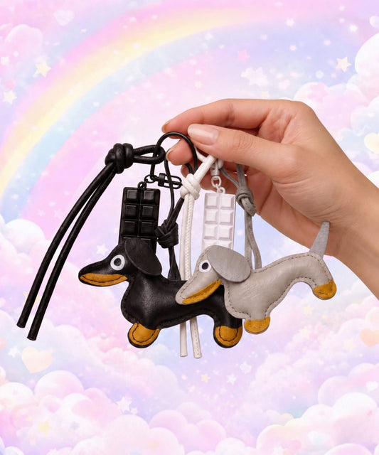 Hand holding black and grey sausage dog PU leather bag charm keychains with chocolate charms against a pastel rainbow background.