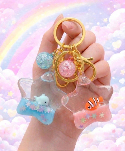 Star liquid glitter keychains with dolphin and clownfish designs held in hand, cute floating ocean animal bag charms with pastel rainbow background
