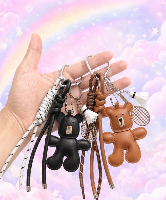 Hand holding black and brown bear PU leather bag charm keychains with badminton racket charms and shuttlecock charms against a pastel rainbow background.