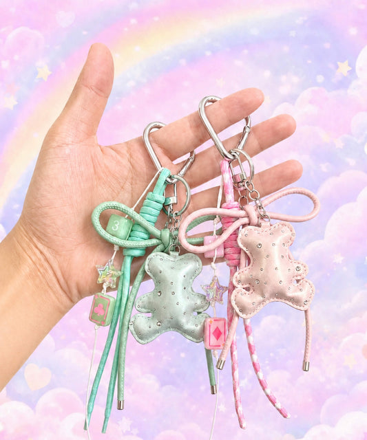 Hand holding green and pink bear PU leather bag charm keychains with sparkle charms, star charms and card charms against a pastel rainbow background.