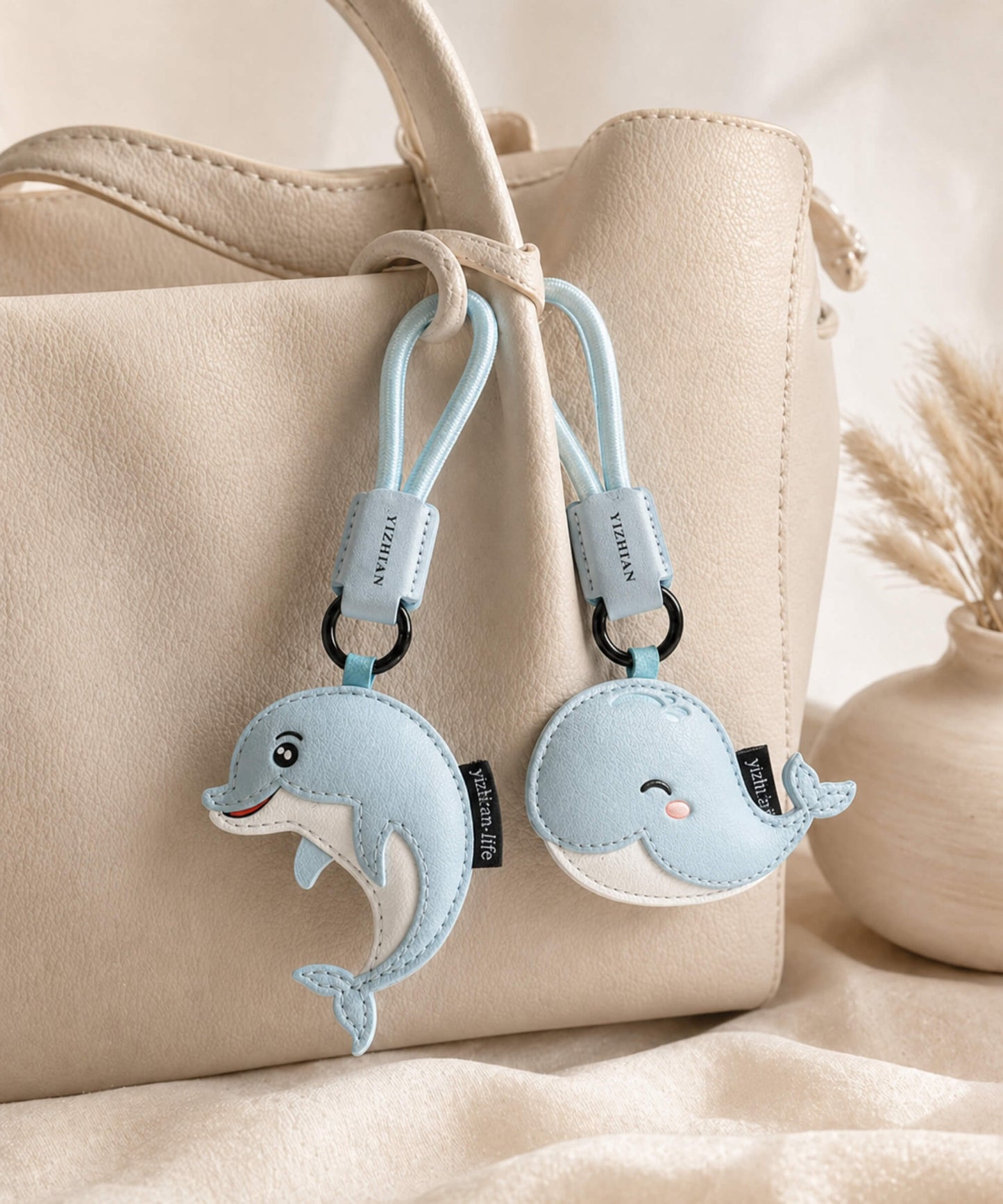 Blue dolphin and whale PU leather bag charms attached to a cream handbag in a soft lifestyle setting.