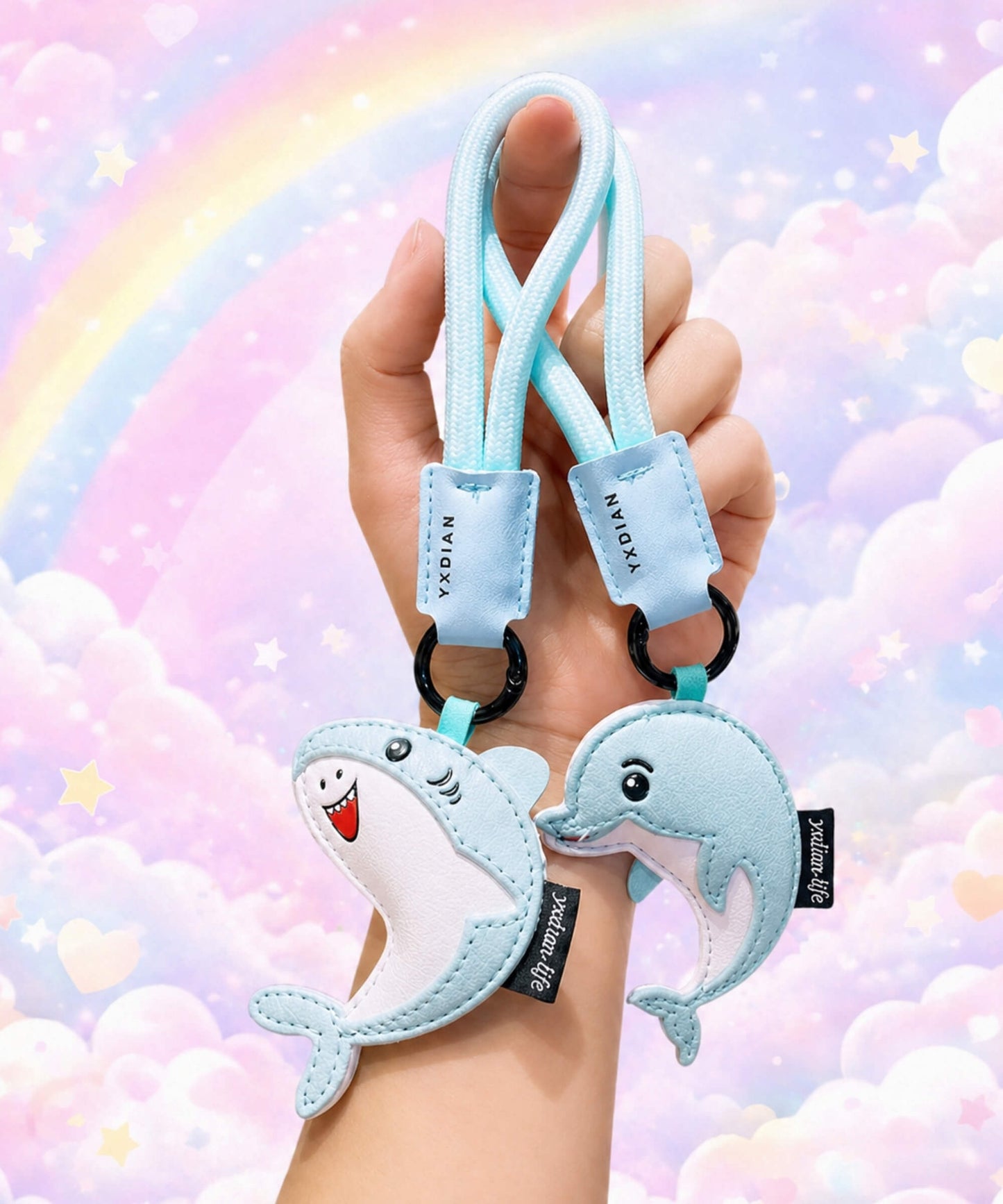 Blue shark and dolphin PU leather keychains held in hand against a pastel rainbow cloud background.