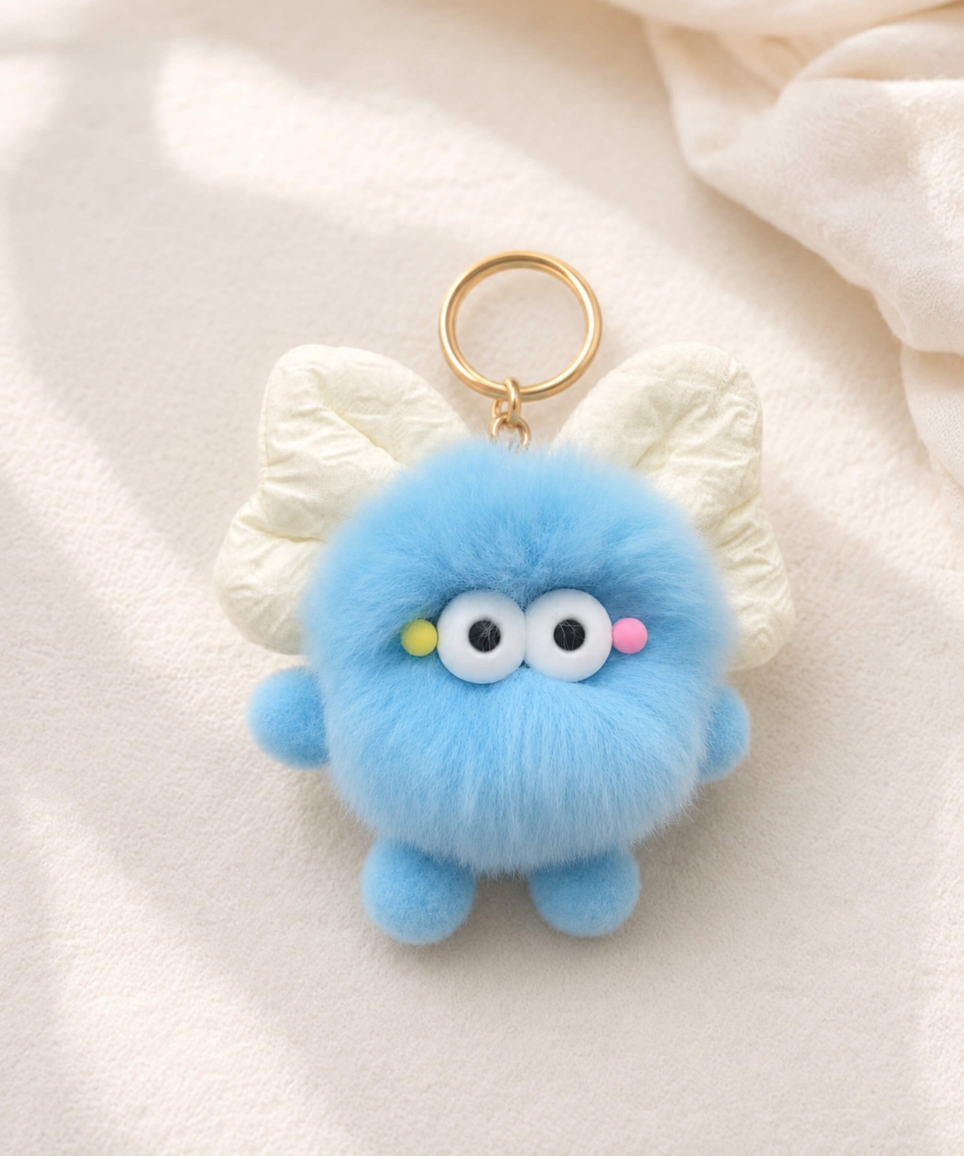 Blue fluffy bow plush keychain on soft cream fabric with cute wide-eye details and gold-tone key ring hardware