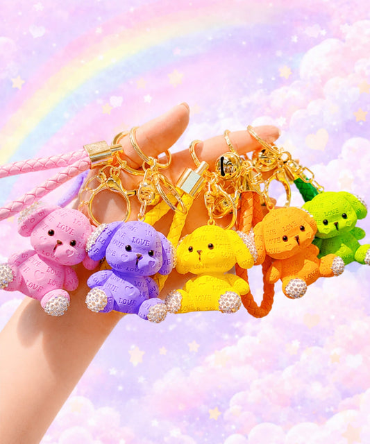 Hand holding pink, purple, yellow, orange and green love puppy resin keychains against a pastel rainbow background.