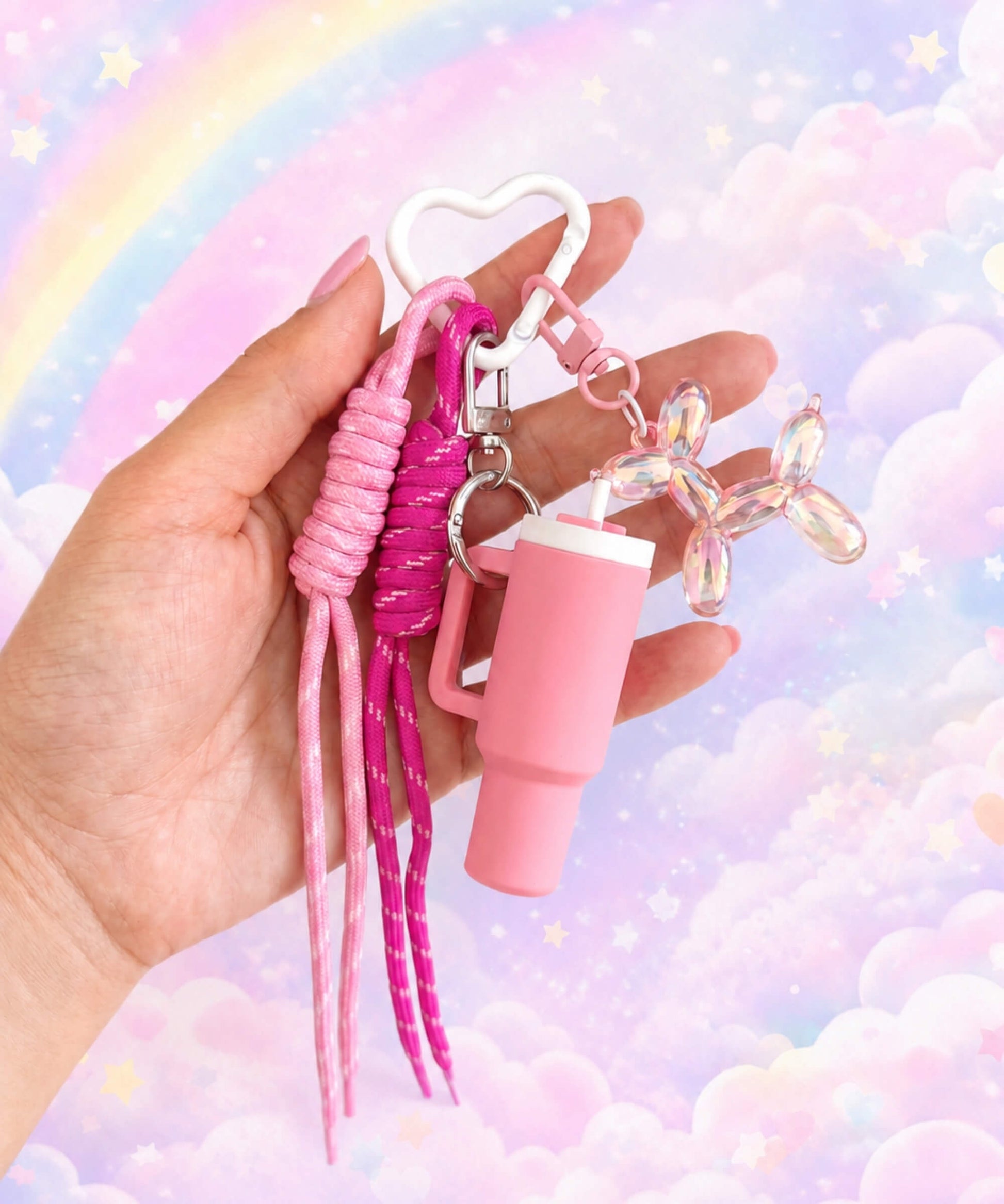 Pink tumbler cup rope keychain held in hand with heart carabiner, rope straps and balloon dog charm on a rainbow cloud background