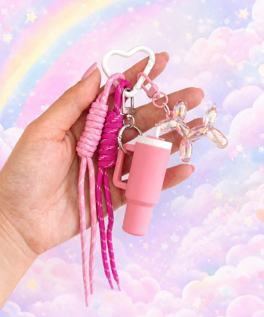 Pink tumbler cup rope keychain held in hand with heart carabiner, rope straps and balloon dog charm on a rainbow cloud background