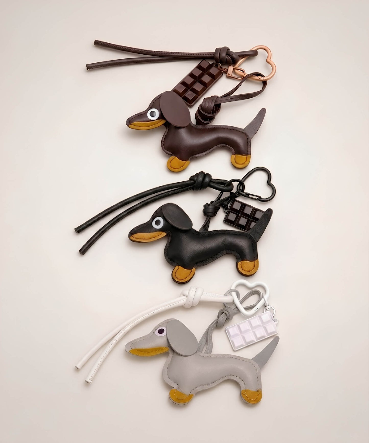 Brown, black and grey sausage dog PU leather bag charm keychains with chocolate charms displayed together on a neutral background.