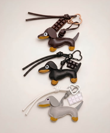 Brown, black and grey sausage dog PU leather bag charm keychains with chocolate charms displayed together on a neutral background.