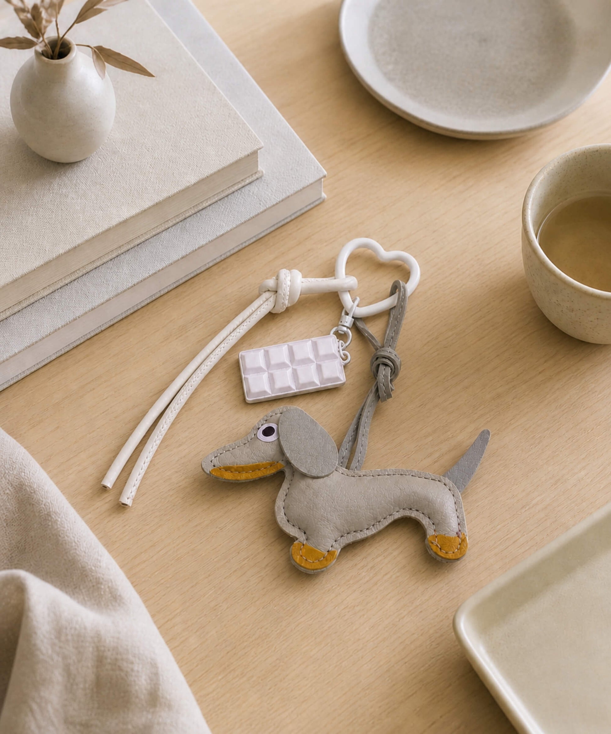 Grey sausage dog PU leather bag charm keychain with a white chocolate charm displayed on a wooden table in a lifestyle setting.