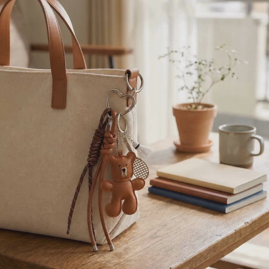 Brown bear PU leather bag charm keychain with a badminton racket charm and shuttlecock charm attached to a tote bag, shown in a lifestyle video with the bag being lifted.