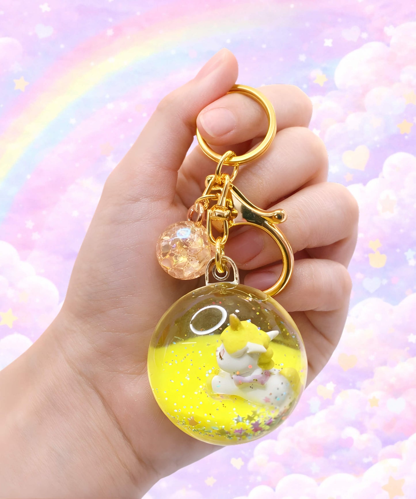Unicorn liquid glitter keychain in yellow colour held in hand, cute floating acrylic bag charm with sparkles and pastel rainbow background